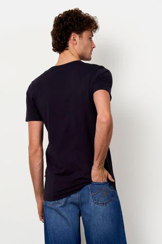 Zwart T-shirt van Le Fabuleux Marcel de Bruxelles, gedragen door een man met donkere krullen, van achteren gezien met een hand in zijn spijkerbroekzak.
