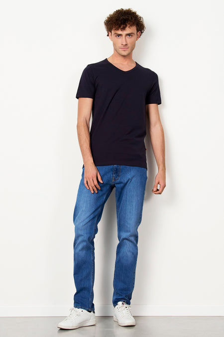 T-shirt noir à col en V et manches courtes de Le Fabuleux Marcel de Bruxelles, porté par un homme avec un jean bleu et des baskets blanches.