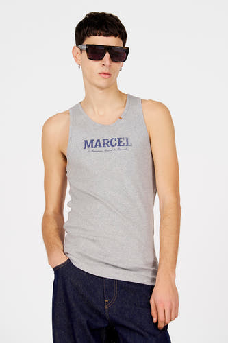 Grijs tanktop van Le Fabuleux Marcel de Bruxelles, met blauwe tekstprint op de borst, gedragen met een donkere jeans en zonnebril.
