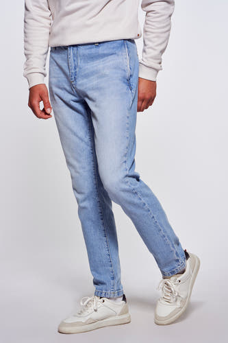 Jeans denim bleu clair 7/8 de Le Fabuleux Marcel de Bruxelles, avec des baskets blanches et un pull clair, debout en vue de profil.