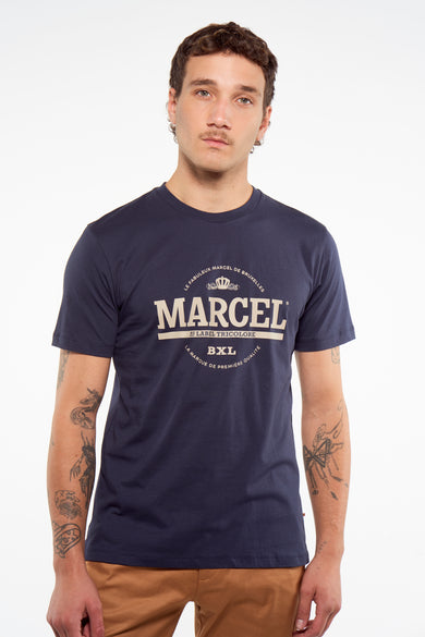 T-shirt bleu marine à manches courtes avec logo blanc de Le Fabuleux Marcel de Bruxelles, porté par un mannequin avec des tatouages et un pantalon marron, vue de face.
