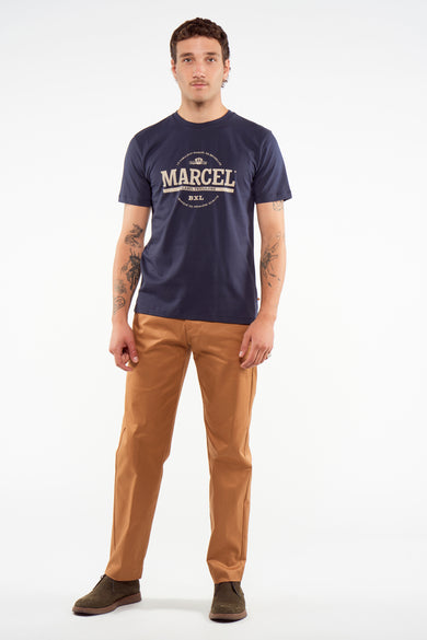 T-shirt bleu marine à manches courtes avec logo Marcel blanc, de Le Fabuleux Marcel de Bruxelles, porté par un mannequin en pantalon marron et chaussures foncées, vue de face