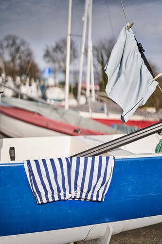 Blauwe zwembroek van Le Fabuleux Marcel de Bruxelles, gestreept, hangend aan een waslijn naast een boot.
