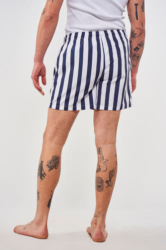 Blauwe zwembroek van Le Fabuleux Marcel de Bruxelles met verticale strepen, gedragen met een wit hemd en tattoos.
