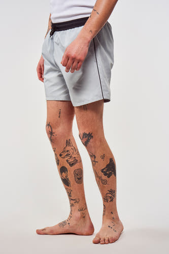 Blauwe zwembroek van Le Fabuleux Marcel de Bruxelles, met zwarte tailleband en tatoeages op de benen, in zij-aanzicht.