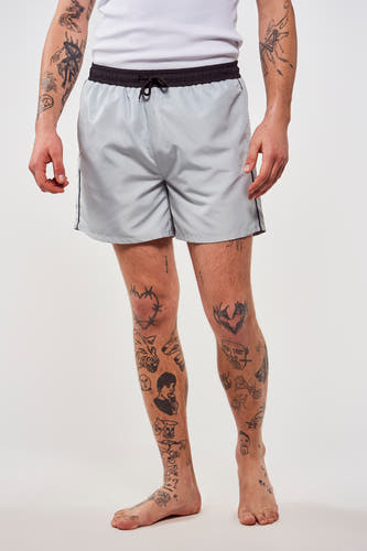 Blauwe zwembroek van Le Fabuleux Marcel de Bruxelles, met zwarte tailleband en tattoos op de benen.
