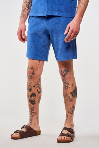 Blauwe short van Le Fabuleux Marcel de Bruxelles, gedragen met bruine sandalen en zichtbare been tatoeages.
