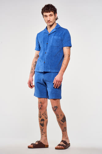 Blauwe short van Le Fabuleux Marcel de Bruxelles, gedragen met een bijpassend overhemd en sandalen.
