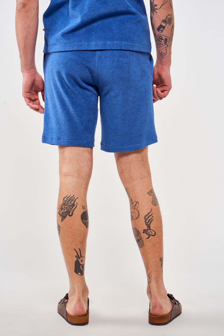 Blauwe short van Le Fabuleux Marcel de Bruxelles, gezien van achteren, met tatoeages op de benen en bruine sandalen.
