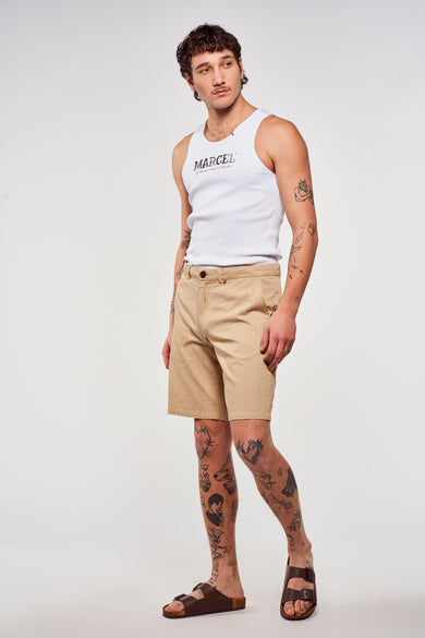 Bruine short van Le Fabuleux Marcel de Bruxelles, gecombineerd met een wit hemd en bruine sandalen, met tatoeages op de benen.
