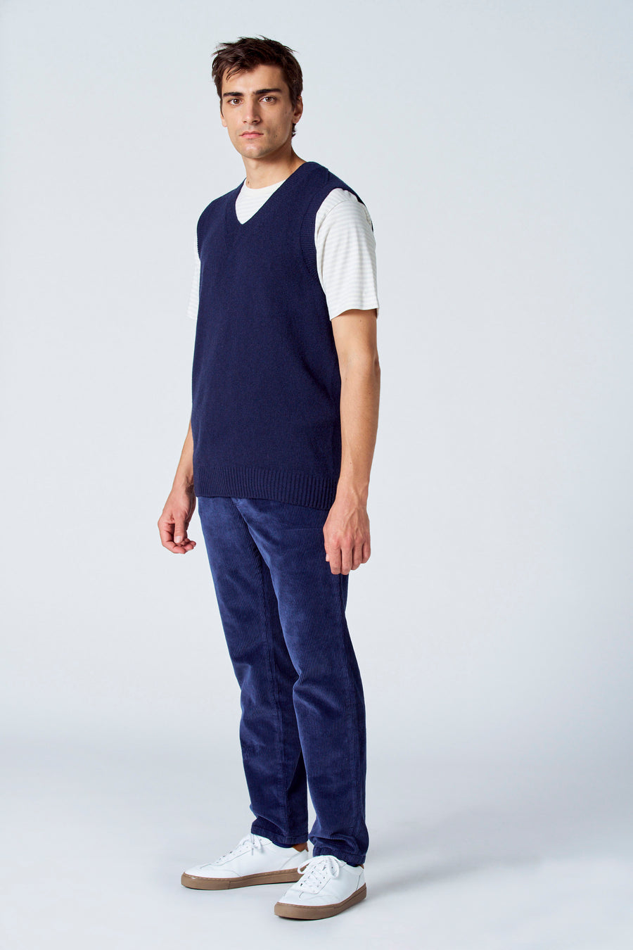 Blauwe debardeur van Le Fabuleux Marcel de Bruxelles, gedragen over een gestreept shirt en corduroy broek, met witte sneakers.