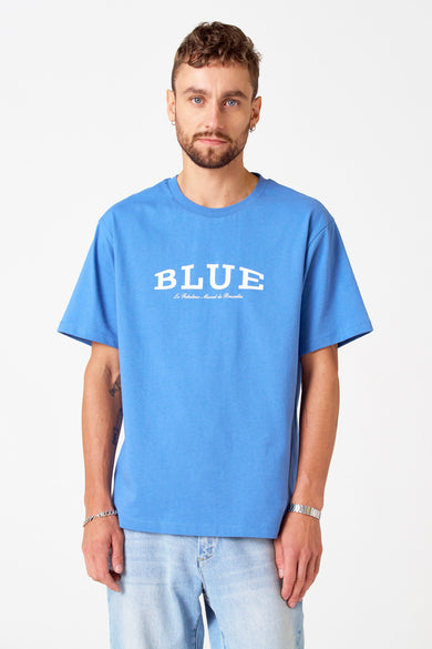 T-shirt bleu à manches courtes de Le Fabuleux Marcel de Bruxelles, porté avec un jean bleu clair et des bracelets en argent.