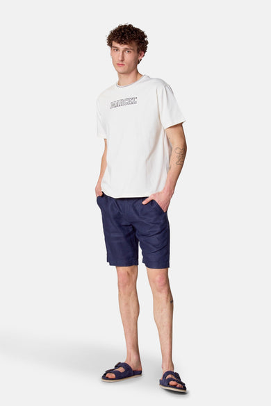 Short bleu de Le Fabuleux Marcel de Bruxelles, porté avec un T-shirt blanc et des sandales bleues.