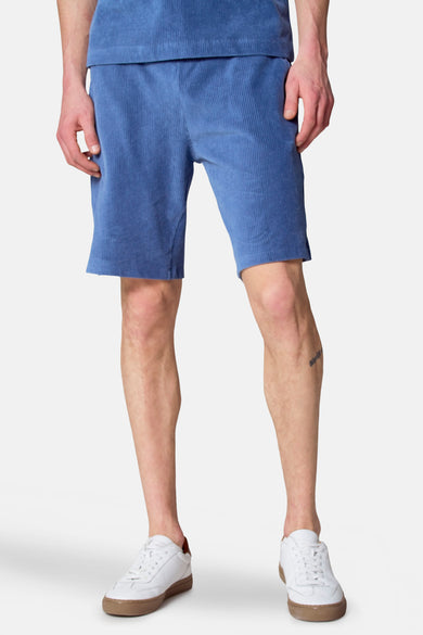 Blauwe corduroy short van Le Fabuleux Marcel de Bruxelles, gedragen met witte sneakers en een blauw shirt.