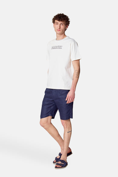 Ecru T-shirt met korte mouwen van Le Fabuleux Marcel de Bruxelles, gedragen met donkerblauwe shorts en sandalen.
