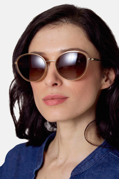 Beige zonnebril van Le Fabuleux Marcel de Bruxelles, met bruine glazen en gouden montuur, gedragen door een vrouw in een denim blouse.
