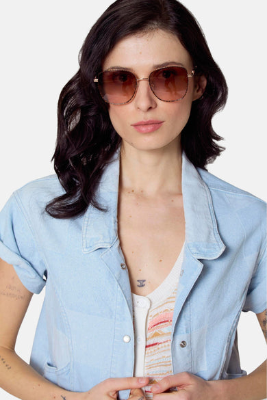 Roze zonnebril van Le Fabuleux Marcel de Bruxelles, met een metalen montuur en bruine glazen, gedragen door een vrouw met een lichtblauwe blouse.