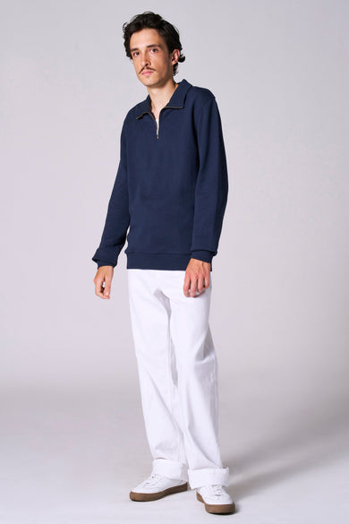 Blauwe sweater van Le Fabuleux Marcel de Bruxelles, gedragen met een witte broek en witte schoenen.
