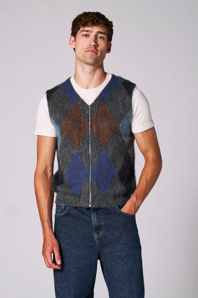 Multikleurig debardeur van Le Fabuleux Marcel de Bruxelles, met ruitpatroon en een ritssluiting, gedragen over een wit T-shirt en een donkere spijkerbroek.
