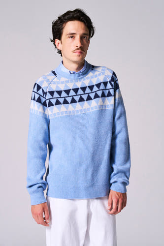 T-shirt bleu avec col rond de Le Fabuleux Marcel de Bruxelles, avec un motif à carreaux sur les épaules, porté avec un pantalon blanc.
