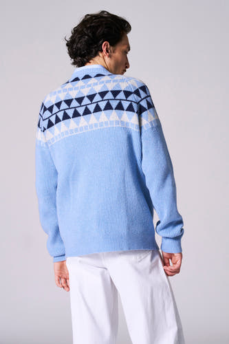 Blauwe trui met ronde hals van Le Fabuleux Marcel de Bruxelles, met een ruitpatroon op de schouders, gedragen met een witte broek.
