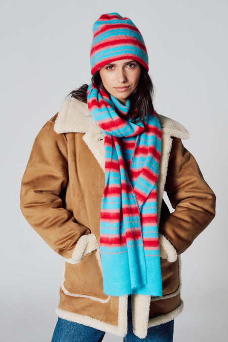 Multicolor wintersjaal van Le Fabuleux Marcel de Bruxelles, met gestreept patroon, gedragen met een bijpassende muts en jas.

