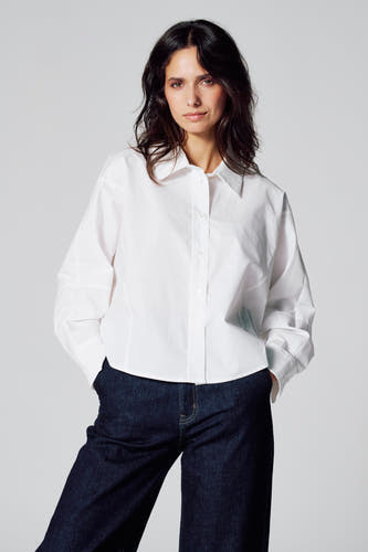 Blouse blanche à manches longues de Le Fabuleux Marcel de Bruxelles, associée à des jeans noirs et les mains dans les poches.