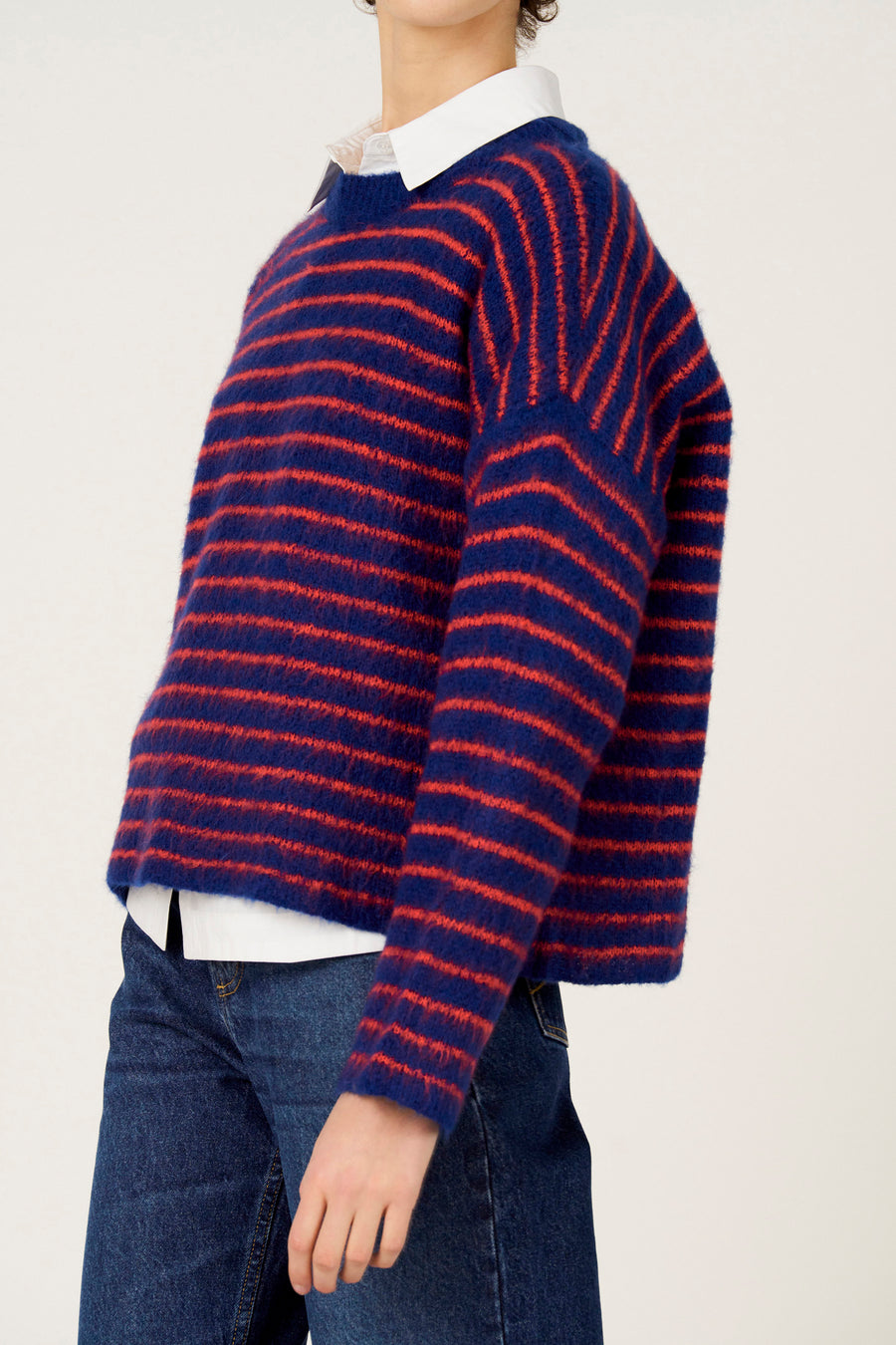 Multikleurige trui met ronde hals van Le Fabuleux Marcel de Bruxelles, met rode strepen, gecombineerd met een wit overhemd en een blauwe jeans.

