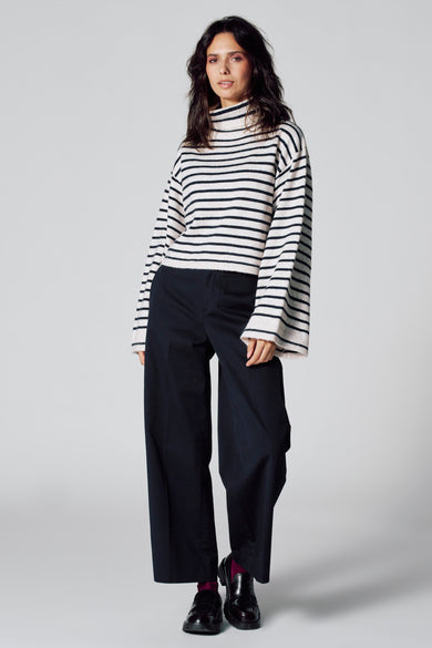 Pull multicolore avec col montant de Le Fabuleux Marcel de Bruxelles, à rayures et manches larges, associé à un pantalon noir et des chaussures.
