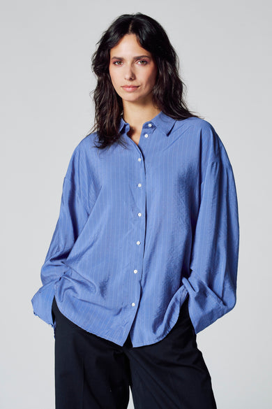 Chemise bleue à rayures horizontales de longues manches de Le Fabuleux Marcel de Bruxelles, avec des boutons blancs et portée avec un pantalon noir.