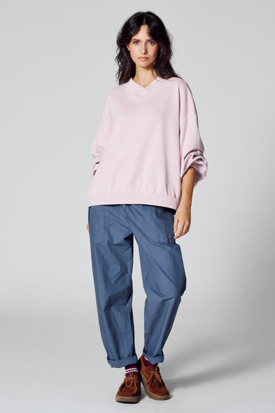 Pull-over rose de Le Fabuleux Marcel de Bruxelles, porté avec un pantalon bleu et des chaussures marrons.