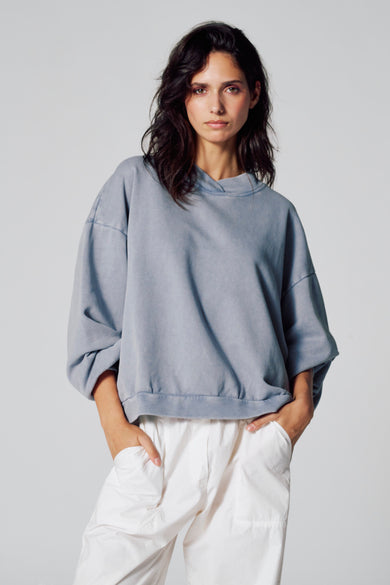 Pull-over bleu avec col rond de Le Fabuleux Marcel de Bruxelles, porté avec un pantalon blanc.
