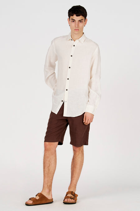 Chemise blanche en lin à manches longues de Le Fabuleux Marcel de Bruxelles, avec boutons foncés, combinée à un short marron.