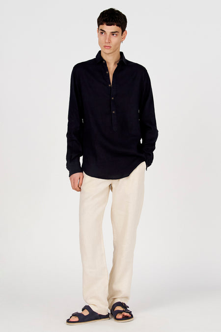 Chemise bleu foncé à manches longues de Le Fabuleux Marcel de Bruxelles, avec col Henley et boutons, portée avec un pantalon beige et des sandales.