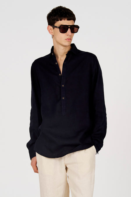 Chemise bleu foncé à manches longues de Le Fabuleux Marcel de Bruxelles, avec une patte de boutonnage courte et des boutons marron, portée avec un pantalon beige et des lunettes de soleil.