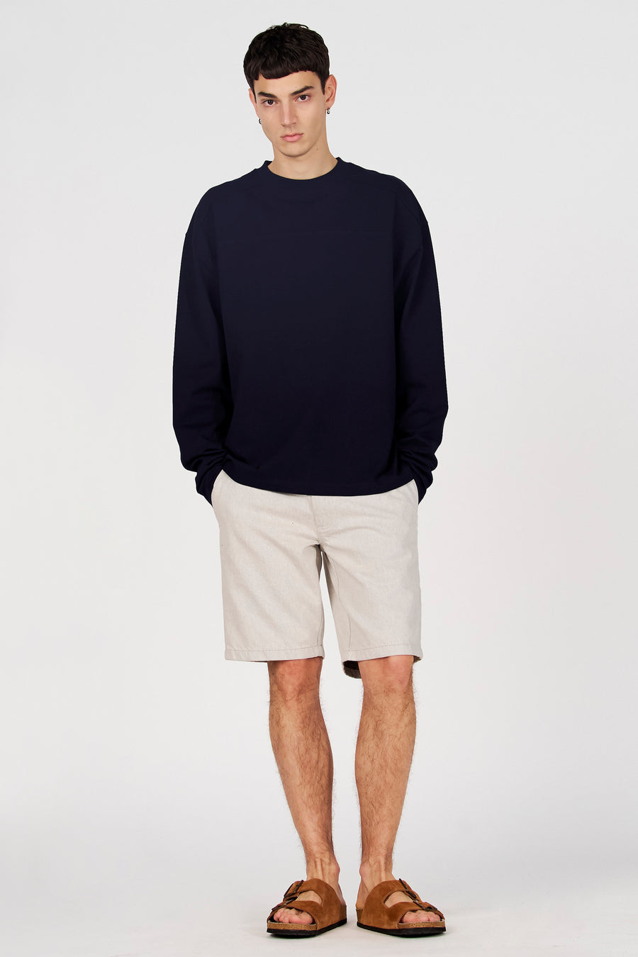 Pull bleu de Le Fabuleux Marcel de Bruxelles, porté avec un short beige et des sandales marron, debout devant un fond blanc.