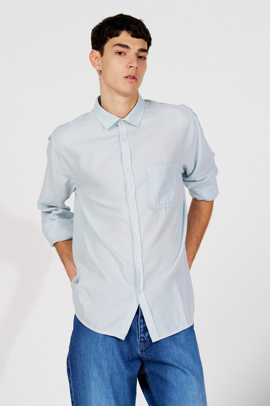 Chemise rayée bleue (manches longues) de Le Fabuleux Marcel de Bruxelles, avec manches retroussées, portée avec un jean bleu foncé.