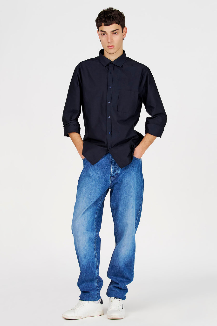 Chemise bleue à manches longues de Le Fabuleux Marcel de Bruxelles, avec fermeture boutonnée et poche poitrine, portée avec un jean bleu et des baskets blanches.