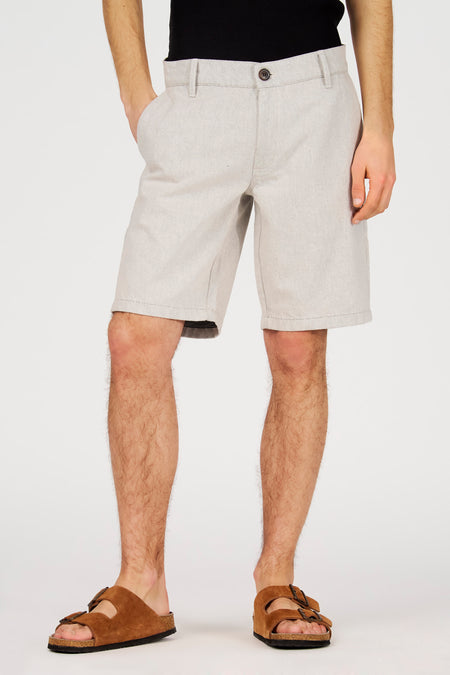 Ecru short van Le Fabuleux Marcel de Bruxelles, met steekzakken en een knoopsluiting, gedragen met bruine sandalen.
