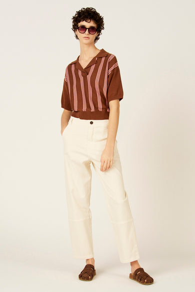 Bruine polo met korte mouwen van Le Fabuleux Marcel de Bruxelles, met roze verticale strepen. Gedragen met crèmekleurige broek en bruine sandalen.