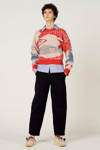 Pull multicolore à col rond de Le Fabuleux Marcel de Bruxelles, avec un motif de poisson et porté sur une chemise rayée avec un pantalon noir.
