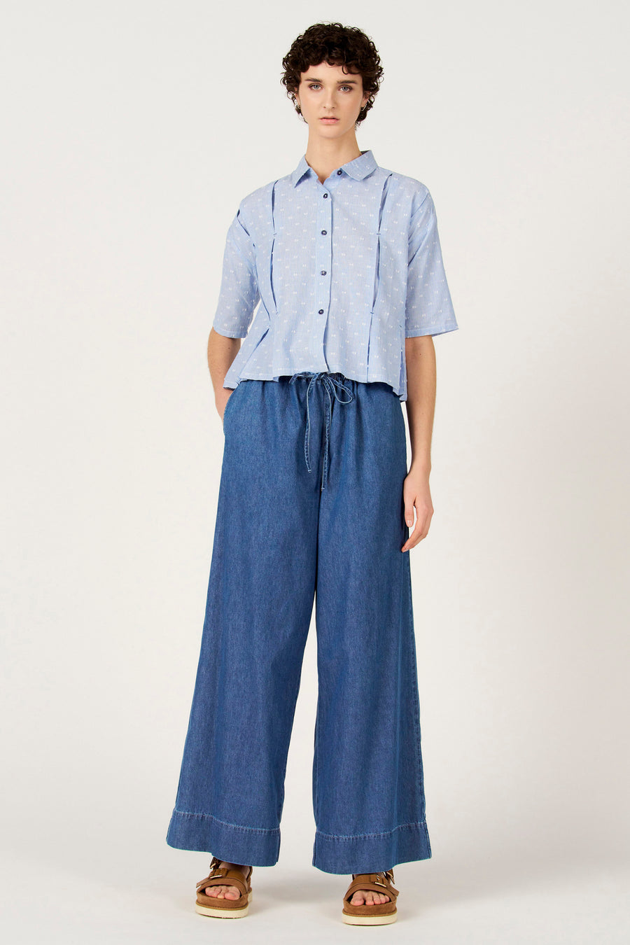 Blauw gestreept overhemd met korte mouwen van Le Fabuleux Marcel de Bruxelles, met witte stippen en gedragen met wijde, donkerblauwe broek.
