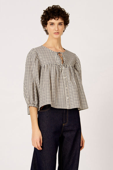 Blauw geruite blouse van Le Fabuleux Marcel de Bruxelles, met driekwart mouwen en een knoopdetail aan de halslijn, gecombineerd met een donkere jeans.
