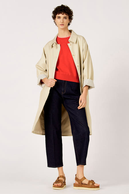 Beige jas van Le Fabuleux Marcel de Bruxelles, gedragen over een rode trui en donkere jeans met bruine sandalen.