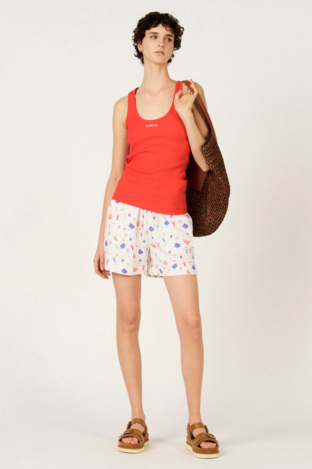 Multikleurige shorts van Le Fabuleux Marcel de Bruxelles, met zomerse print van strandtaferelen, gedragen met een rode top en bruine sandaaltjes.