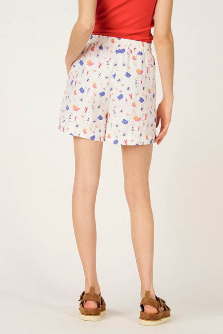 Multikleurige korte broek van Le Fabuleux Marcel de Bruxelles, met strandthema-patroon, gecombineerd met rode top en bruine sandalen.
