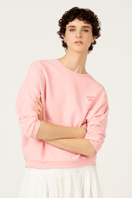 Roze sweater met ronde hals van Le Fabuleux Marcel de Bruxelles, met kleine rode opdruk en witte geplooide rok.
