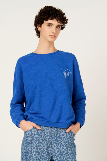 Blauwe sweater met ronde hals van Le Fabuleux Marcel de Bruxelles, met borduursel op de borst en blauw-bloemige broek.
