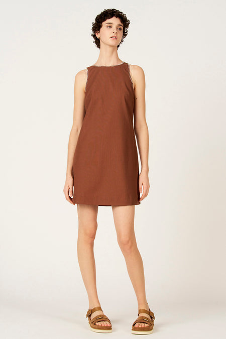 Bruine mouwloze kleedje van Le Fabuleux Marcel de Bruxelles, met witte sierstiksels langs de halslijn en armsgaten, gecombineerd met bruine sandalen.
