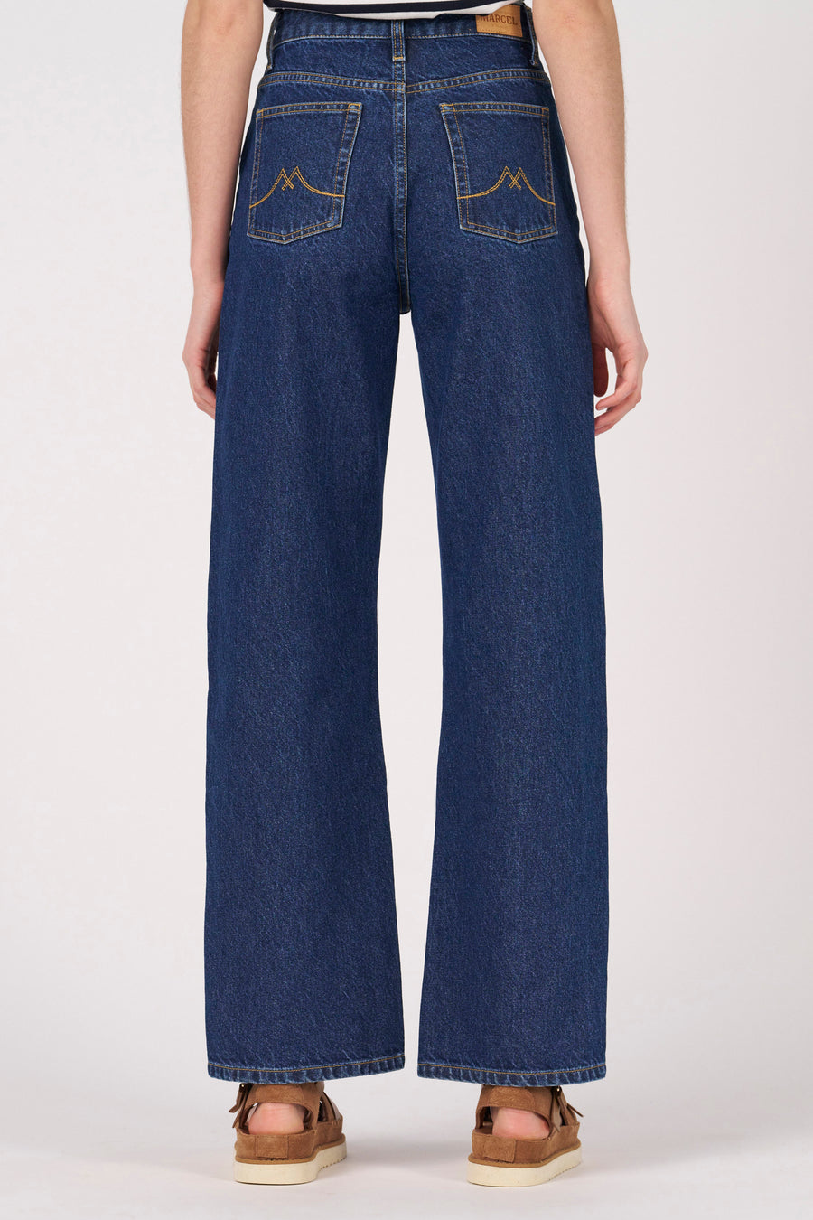 Donkerblauwe wijde jeans van Le Fabuleux Marcel de Bruxelles, met opgestikte zakken aan de achterkant, gedragen met bruine sandalen.
